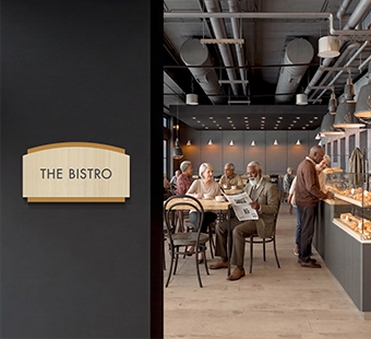 A group of seniors enjoys socializing on an independent living caf&eacute;. The space is marked by a Flexia Pinnacle 2&trade; room sign.