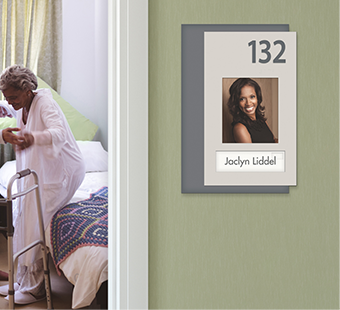 A nurse helps a senior stand inside her room. Outside, an Innovate patient room number sign with photo helps the resident find her room with ease.