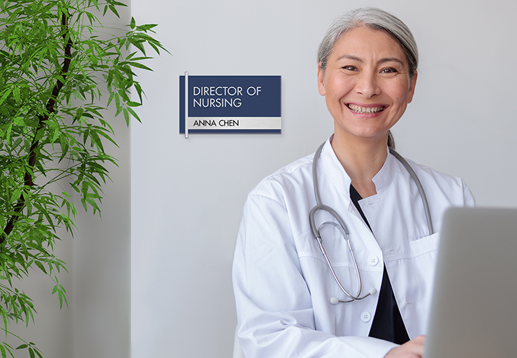 A nurse wearing a lab coat smiles from the nurse's station. Behind her is a NURSE ON DUTY sign with nameplate insert.