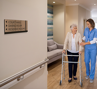 A nurse assists a rehab patient with a walker. Clear wayfinding help ensure patients can navigate independently as rehabilitation progresses.