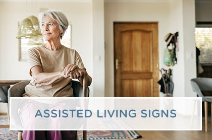 A female resident sits with a walking cane in a brightly lit, comfortable assisted living suite.