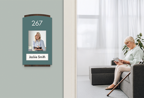 Resident room sign with photo insert beside an older woman seated in her memory care living room