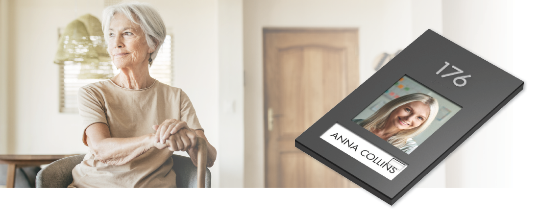 Older woman seated in an assisted living room beside a personalized resident room sign with picture