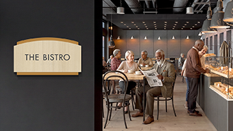A group of seniors enjoys socializing on an independent living caf&eacute;. The space is marked by a Flexia Pinnacle 2&trade; room sign.