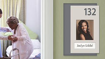 A nurse helps a senior stand inside her room. Outside, an Innovate patient room number sign with photo helps the resident find her room with ease.