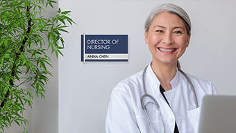 A nurse wearing a lab coat smiles from the nurse's station. Behind her is a NURSE ON DUTY sign with nameplate insert.