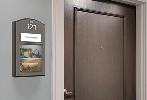 A resident walks through a modern senior care corridor with clear room identification and accessible wayfinding signs.
