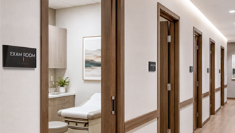 A modern medical office corridor with Facets woodgrain exam room signs mounted on the latch side of each exam room door.