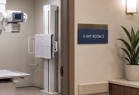 X-Ray Room 3 sign mounted outside an outpatient radiology room with visible x-ray table, overhead imaging equipment, and wall bucky inside.