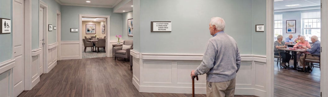 A resident walks through a modern senior care corridor with clear room identification and accessible wayfinding signs.