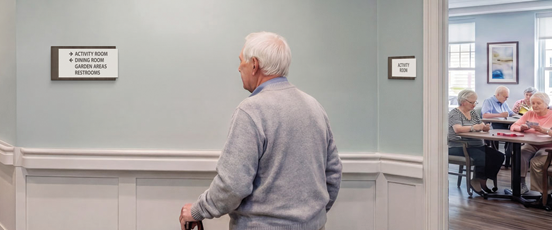 A resident walks through a modern senior care corridor with clear room identification and accessible wayfinding signs.